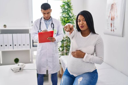Téléchargez les photos : Jeune mère hispanique attendant un bébé chez le médecin pointant du doigt vers la caméra et vers vous, geste confiant et sérieux - en image libre de droit