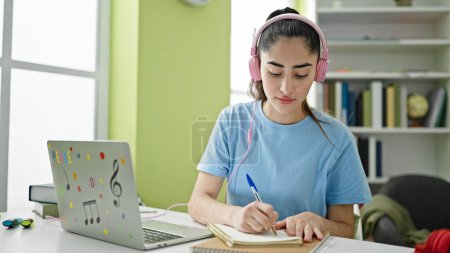 Téléchargez les photos : Jeune belle étudiante hispanique utilisant ordinateur portable et écouteurs prenant des notes à l'université de la bibliothèque - en image libre de droit