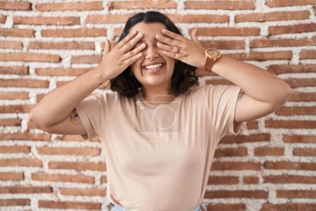 Foto de Mujer hispana joven de pie sobre la pared de ladrillos cubriendo los ojos con las manos sonriendo alegre y divertido. concepto ciego. - Imagen libre de derechos