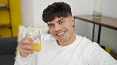 Foto de Joven hombre hispano sonriendo confiado sosteniendo un vaso de jugo en casa - Imagen libre de derechos