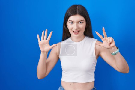 Foto de Joven mujer caucásica de pie sobre fondo azul mostrando y señalando hacia arriba con los dedos número ocho mientras sonríe confiado y feliz. - Imagen libre de derechos