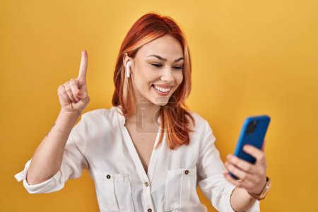 Photo for Young caucasian woman using smartphone wearing earphones smiling with an idea or question pointing finger with happy face, number one - Royalty Free Image