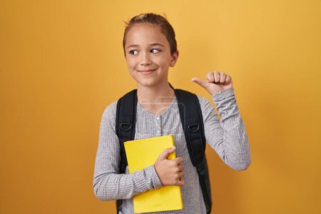 Téléchargez les photos : Petit garçon caucasien portant un sac à dos étudiant et tenant un livre pointant le pouce vers le côté souriant heureux avec la bouche ouverte - en image libre de droit