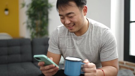 Téléchargez les photos : Jeune homme chinois utilisant smartphone boire du café à la maison - en image libre de droit