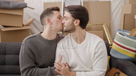 Photo for Two men couple sitting on sofa hugging each other kissing at new home - Royalty Free Image