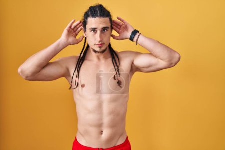 Photo for Hispanic man with long hair standing shirtless over yellow background trying to hear both hands on ear gesture, curious for gossip. hearing problem, deaf - Royalty Free Image