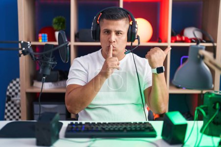 Téléchargez les photos : Jeune homme hispanique jouant à des jeux vidéo demandant à être silencieux avec le doigt sur les lèvres pointant avec la main sur le côté. silence et concept secret. - en image libre de droit