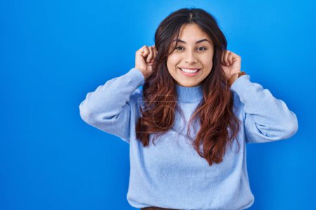 Téléchargez les photos : Jeune femme hispanique debout sur fond bleu souriant tirant les oreilles avec les doigts, geste drôle. problème d'audition - en image libre de droit
