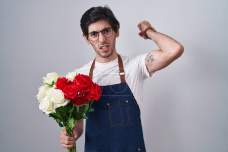 Téléchargez les photos : Jeune homme hispanique tenant un bouquet de roses blanches et rouges en colère et fou levant le poing frustré et furieux tout en criant de colère. rage et concept agressif. - en image libre de droit