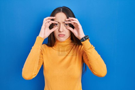 Photo for Young brunette woman standing over blue background trying to open eyes with fingers, sleepy and tired for morning fatigue - Royalty Free Image