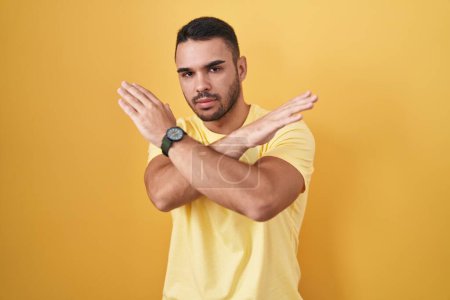 Foto de Joven hombre hispano de pie sobre fondo amarillo expresión de rechazo cruzando brazos haciendo signo negativo, cara enojada - Imagen libre de derechos
