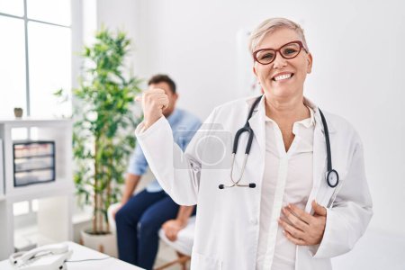 Téléchargez les photos : Médecin féminin portant uniforme et stéthoscope à la clinique médicale pointant le pouce vers le côté souriant heureux avec la bouche ouverte - en image libre de droit