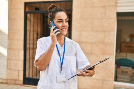 Téléchargez les photos : Jeune belle hispanique femme physiothérapeute lecture document parler sur smartphone à la rue - en image libre de droit
