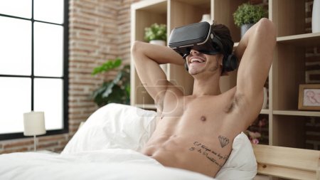 Téléchargez les photos : Jeune homme hispanique assis sur le lit en utilisant des lunettes de réalité virtuelle dans la chambre - en image libre de droit