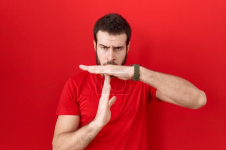 Foto de Joven hombre hispano vistiendo camiseta roja casual haciendo un gesto de tiempo fuera con las manos, rostro frustrado y serio - Imagen libre de derechos