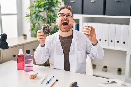 Photo for Middle age caucasian dentist man holding denture and doughnuts angry and mad screaming frustrated and furious, shouting with anger looking up. - Royalty Free Image