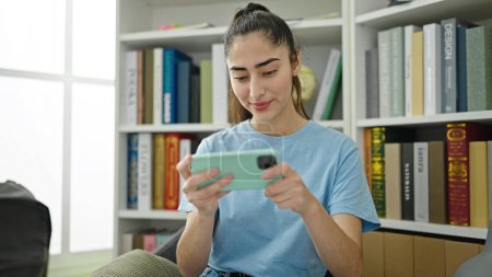 Téléchargez les photos : Jeune belle étudiante hispanique regardant une vidéo sur smartphone à la bibliothèque de l'université - en image libre de droit