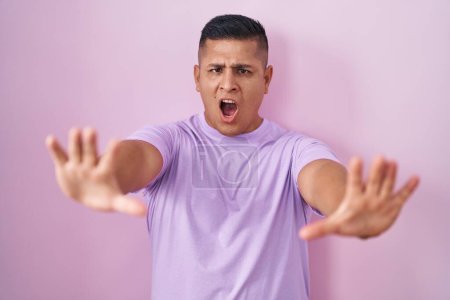 Photo for Young hispanic man standing over pink background doing stop gesture with hands palms, angry and frustration expression - Royalty Free Image