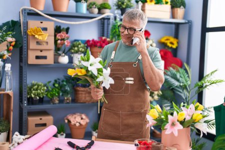 Téléchargez les photos : Moyen Age homme aux cheveux gris fleuriste parler sur smartphone tenant des fleurs à la boutique de fleurs - en image libre de droit