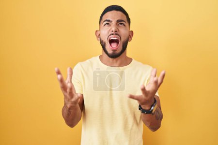 Foto de Young hispanic man standing over yellow background crazy and mad shouting and yelling with aggressive expression and arms raised. frustration concept. - Imagen libre de derechos