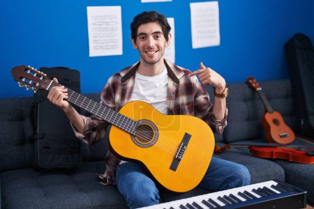 Foto de Joven hombre hispano tocando la guitarra clásica en el estudio de música señalando con el dedo a uno mismo sonriendo feliz y orgulloso - Imagen libre de derechos