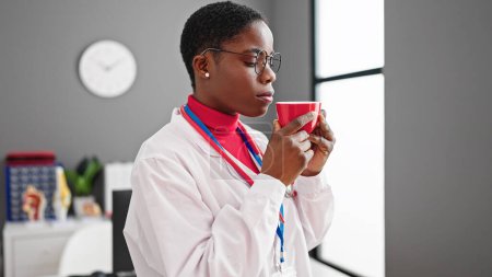 Photo for African american woman doctor smelling cup of coffee at clinic - Royalty Free Image