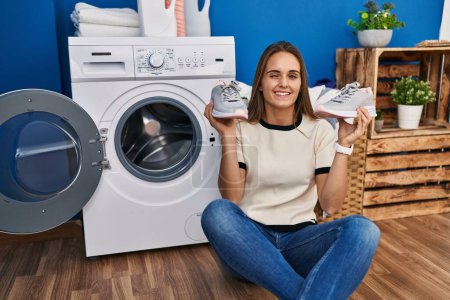 Photo for Young woman putting sneakers in washing machine winking looking at the camera with sexy expression, cheerful and happy face. - Royalty Free Image