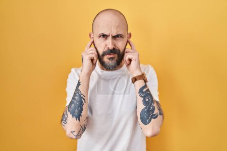 Téléchargez les photos : Young hispanic man with tattoos standing over yellow background covering ears with fingers with annoyed expression for the noise of loud music. deaf concept. - en image libre de droit