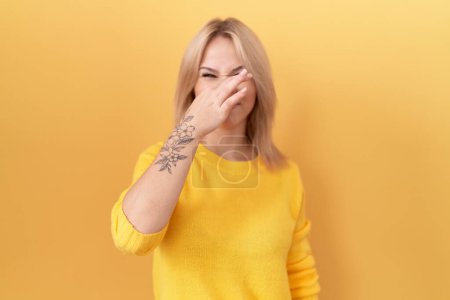 Téléchargez les photos : Jeune femme caucasienne portant un pull jaune sentant quelque chose de puant et dégoûtant, odeur intolérable, retenant son souffle avec les doigts sur le nez. mauvaise odeur - en image libre de droit