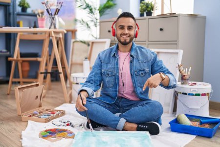 Téléchargez les photos : Jeune homme hispanique peintre assis sur le sol à l'atelier d'art souriant heureux pointant avec la main et le doigt - en image libre de droit