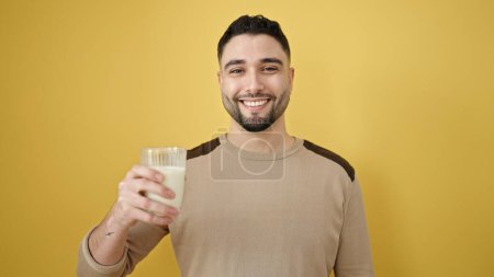 Téléchargez les photos : Jeune homme arabe souriant confiant tenant verre de lait sur fond jaune isolé - en image libre de droit