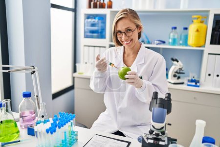 Foto de Mujer joven rubia científica inyectando líquido en manzana en el laboratorio - Imagen libre de derechos