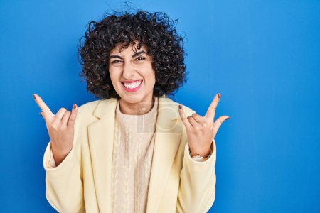 Photo for Young brunette woman with curly hair standing over blue background shouting with crazy expression doing rock symbol with hands up. music star. heavy music concept. - Royalty Free Image