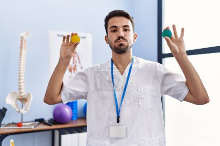 Foto de Joven fisioterapeuta hispano hombre sosteniendo bolas de fuerza para entrenar los músculos de la mano hinchando mejillas con cara divertida. boca inflada con aire, captación de aire. - Imagen libre de derechos