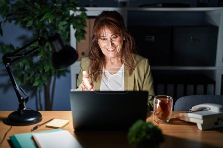 Photo for Middle age hispanic woman working using computer laptop at night smiling friendly offering handshake as greeting and welcoming. successful business. - Royalty Free Image