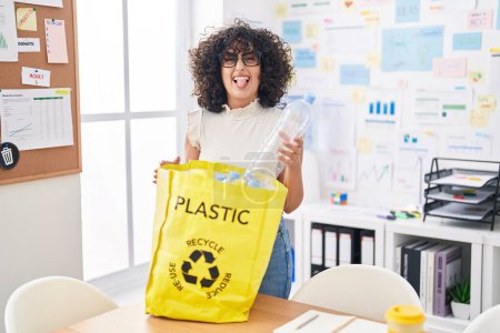 Téléchargez les photos : Jeune femme du Moyen-Orient tenant sac de recyclage avec des bouteilles en plastique au bureau langue collante heureux avec une expression drôle. - en image libre de droit