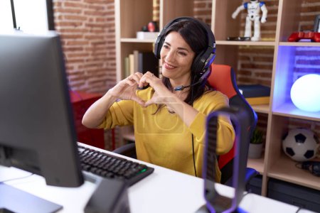 Téléchargez les photos : Femme hispanique du Moyen Âge jouant à des jeux vidéo à l'aide d'écouteurs souriant dans l'amour montrant le symbole du cœur et la forme avec les mains. concept romantique. - en image libre de droit