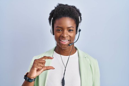Foto de Mujer afroamericana que usa auriculares de agente de centro de llamadas sonriendo y con gesto seguro con la mano haciendo letrero de tamaño pequeño con los dedos mirando y la cámara. concepto de medida. - Imagen libre de derechos