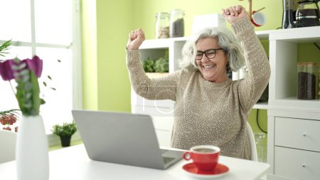 Téléchargez les photos : Femme d'âge moyen avec les cheveux gris en utilisant un ordinateur portable assis sur la table dansant à la maison - en image libre de droit