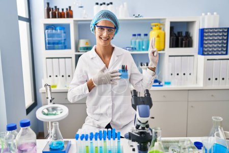 Téléchargez les photos : Femme brune travaillant au laboratoire scientifique souriant jurant avec la main sur la poitrine et les doigts vers le haut, faisant un serment de fidélité - en image libre de droit