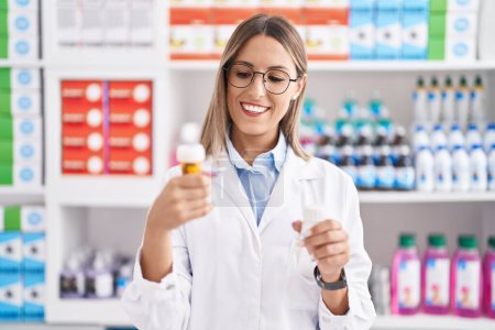 Téléchargez les photos : Jeune belle hispanique femme pharmacien souriant confiant tenant bouteilles pilules à la pharmacie - en image libre de droit