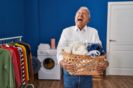 Foto de Hombre mayor con el pelo gris sosteniendo la cesta de la ropa en casa enojado y loco gritando frustrado y furioso, gritando con ira mirando hacia arriba. - Imagen libre de derechos