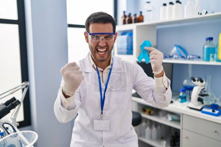 Téléchargez les photos : Jeune homme hispanique avec barbe travaillant au laboratoire scientifique tenant un ruban bleu ennuyé et frustré criant de colère, criant fou de colère et la main levée - en image libre de droit