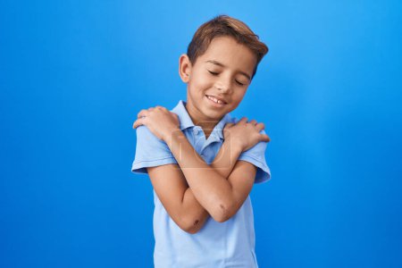 Photo for Little hispanic boy wearing casual blue t shirt hugging oneself happy and positive, smiling confident. self love and self care - Royalty Free Image