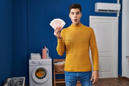 Photo for Young hispanic man at laundry room holding shekels scared and amazed with open mouth for surprise, disbelief face - Royalty Free Image