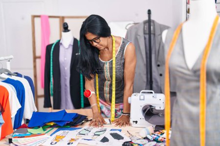 Téléchargez les photos : Femme hispanique du Moyen Âge tailleur regardant la conception de vêtements au magasin de tailleur - en image libre de droit