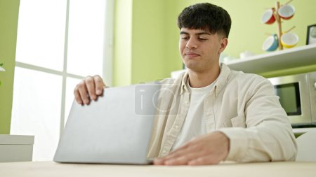 Téléchargez les photos : Jeune homme hispanique ouverture ordinateur portable assis sur la table à la salle à manger - en image libre de droit