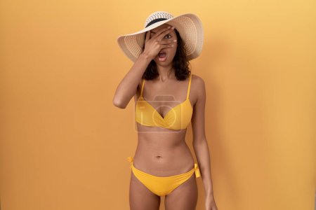 Foto de Mujer hispana joven con bikini y sombrero de verano asomándose en shock cubriendo la cara y los ojos con la mano, mirando a través de los dedos con expresión avergonzada. - Imagen libre de derechos
