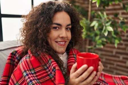 Téléchargez les photos : Young beautiful hispanic woman drinking coffee covering with blanket at home - en image libre de droit