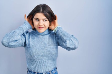 Foto de Mujer hispana joven de pie sobre fondo azul tratando de escuchar ambos gestos de las manos en el oído, curioso por chismes. problema auditivo, sordo - Imagen libre de derechos